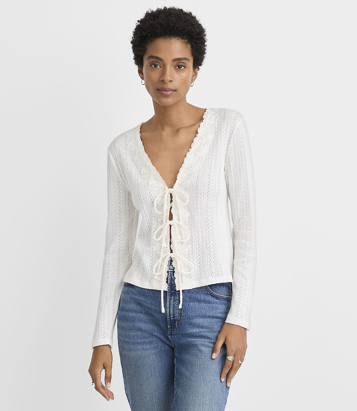A woman in a white lace-up top and blue jeans stands against a plain white background.