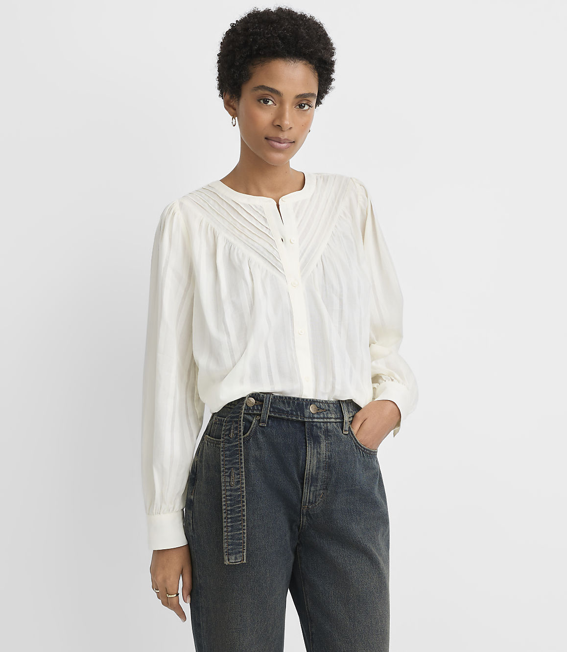 A woman in a white blouse and dark jeans stands against a plain white background.