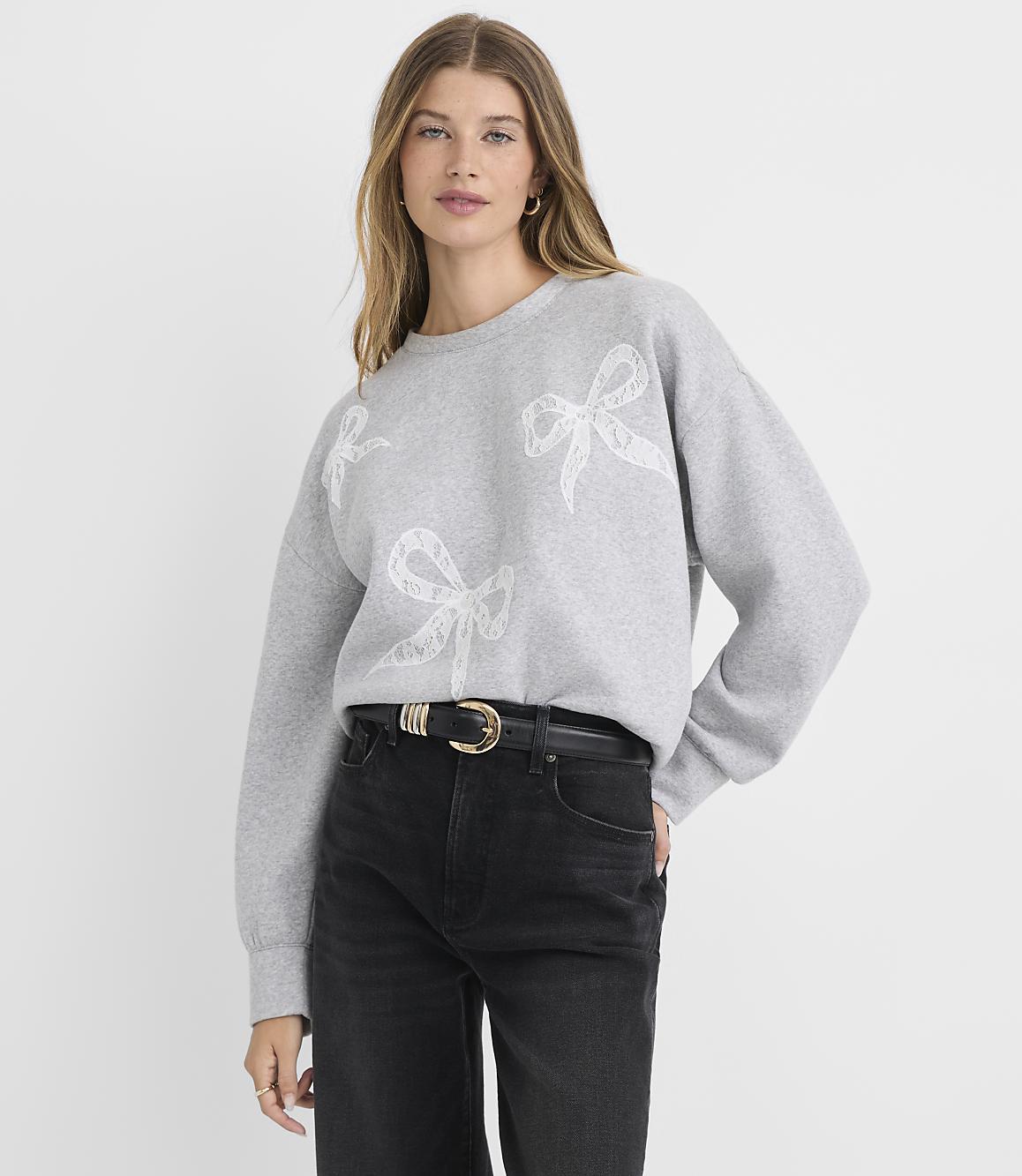A woman in a grey sweatshirt with bow designs and black jeans stands against a plain grey background.