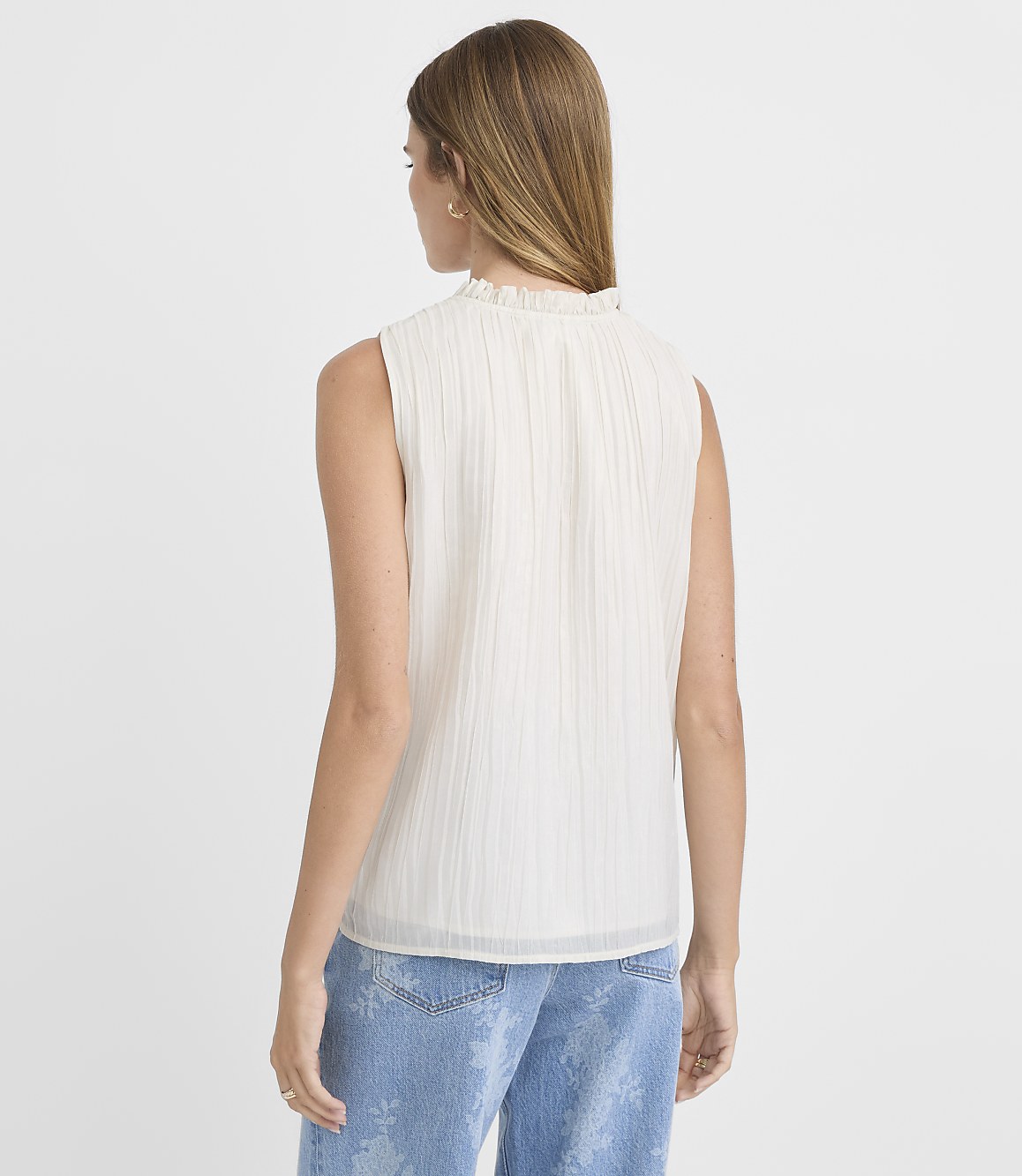 A woman in a white ruffled-neck sleeveless top and distressed jeans stands against a plain white background.