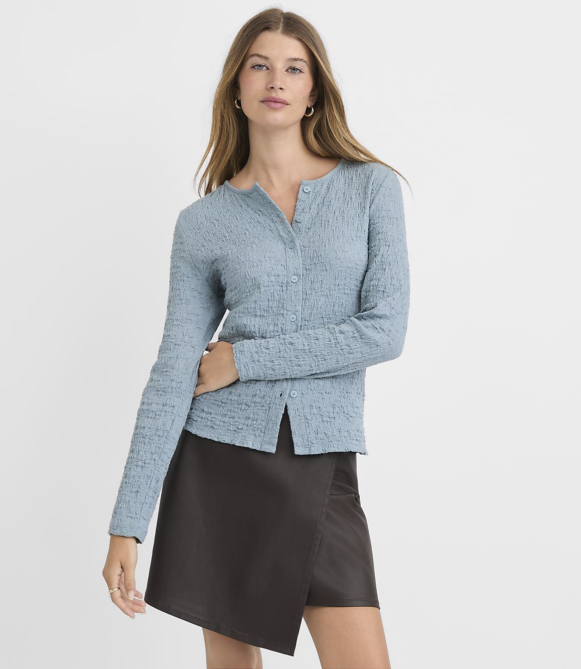 A woman in a textured blue cardigan and a dark brown skirt stands against a plain, light gray background.