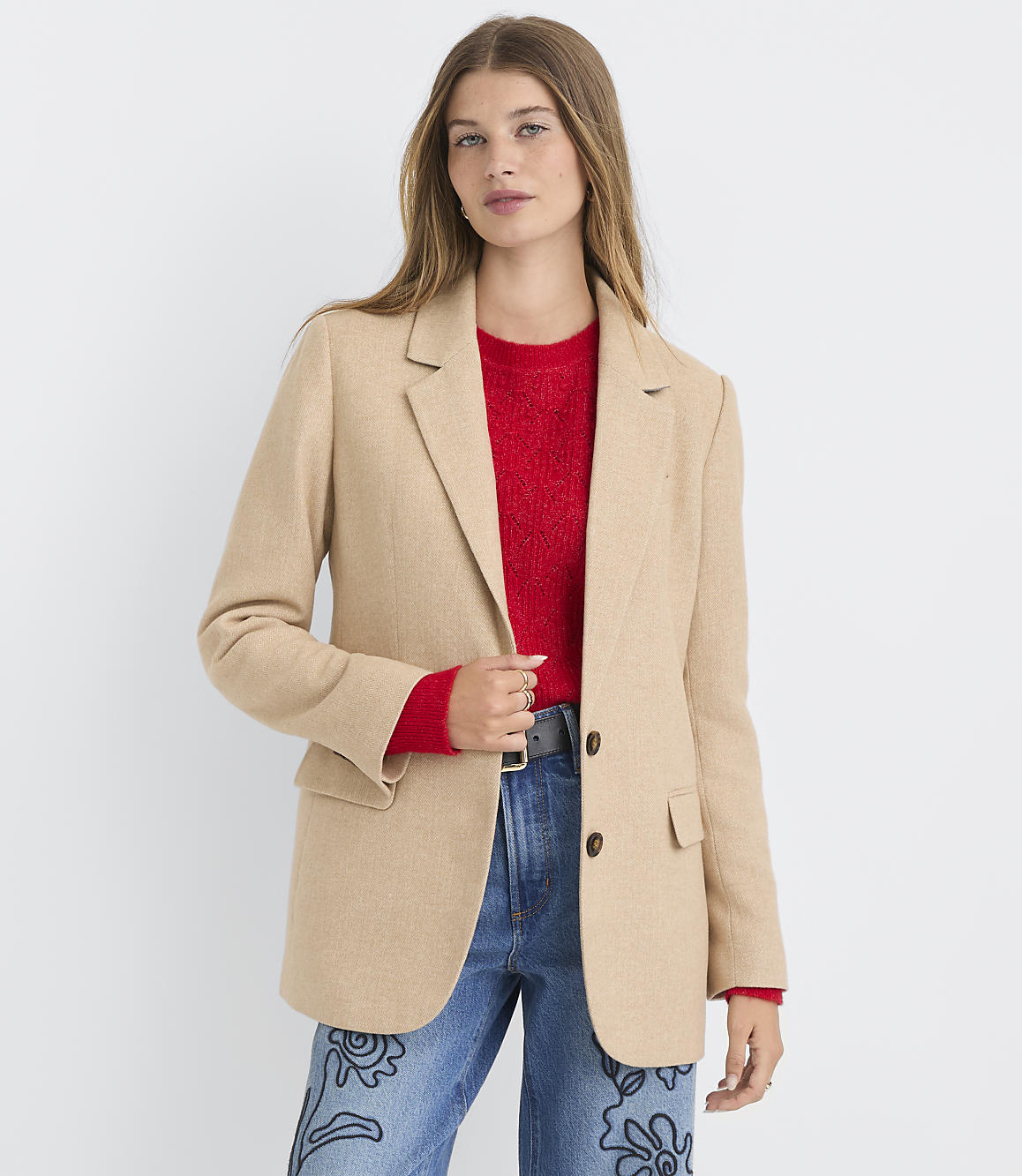 A woman in a tan blazer and red sweater stands against a plain white background.
