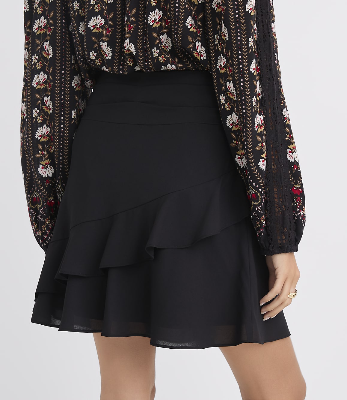 A woman in a black ruffled skirt and a floral print blouse stands against a plain white background.