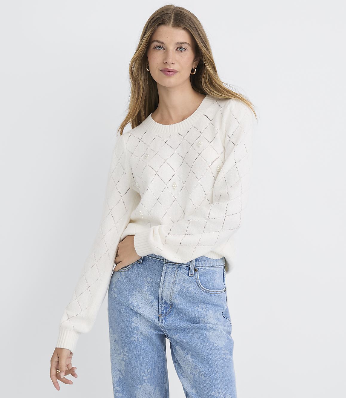 A woman in a cream-colored argyle sweater and light blue patterned jeans stands against a plain white background.