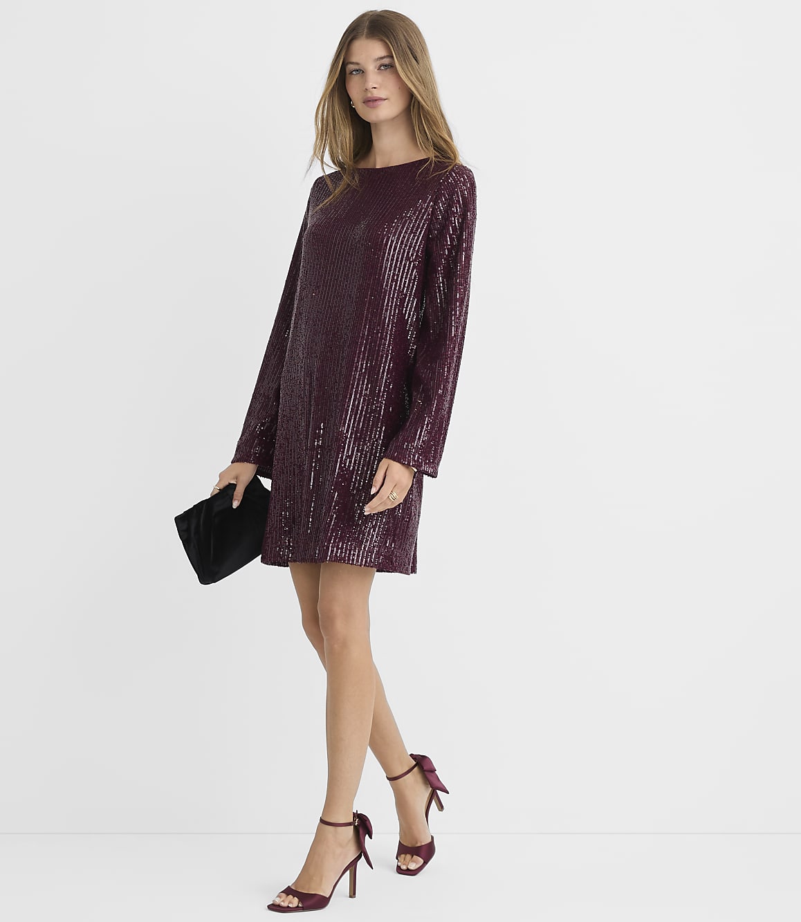 A woman in a burgundy sequined dress and heels holds a black clutch in front of a plain white background.