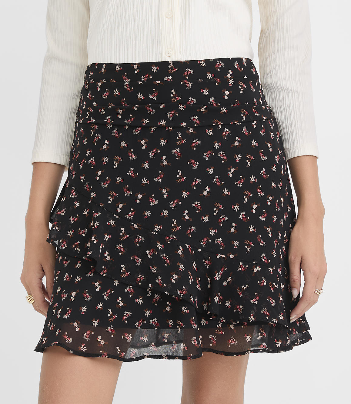 A woman in a black floral mini skirt with ruffles stands against a plain white background.