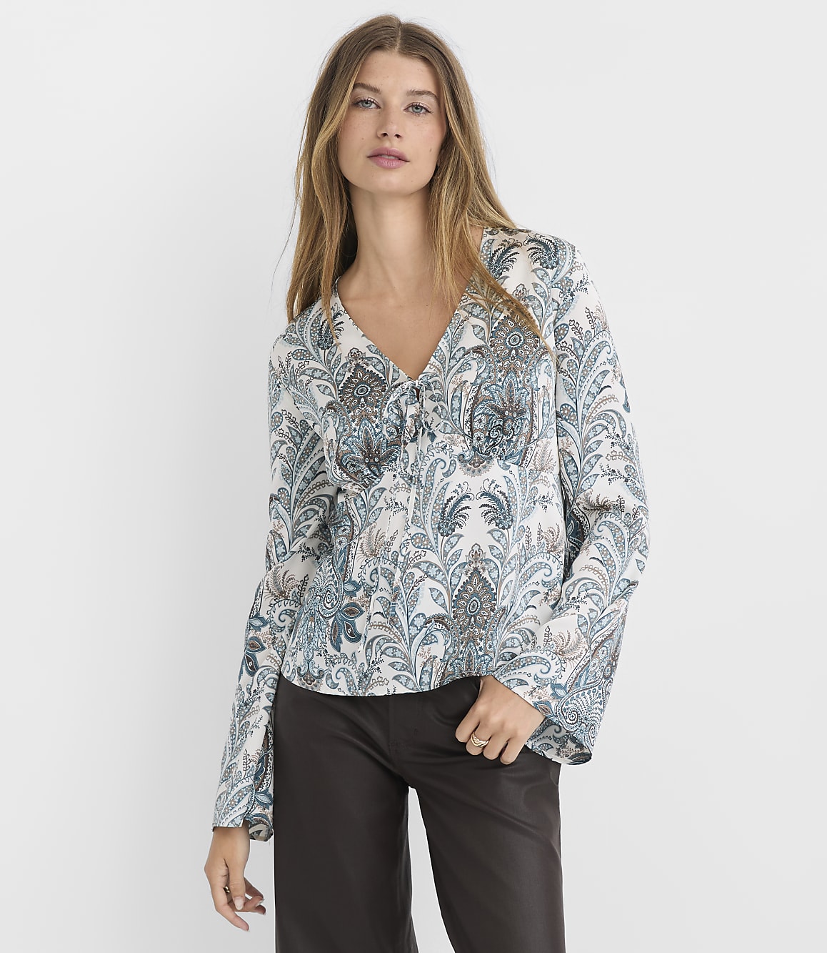 A woman in a patterned blouse and dark pants stands against a plain white background.
