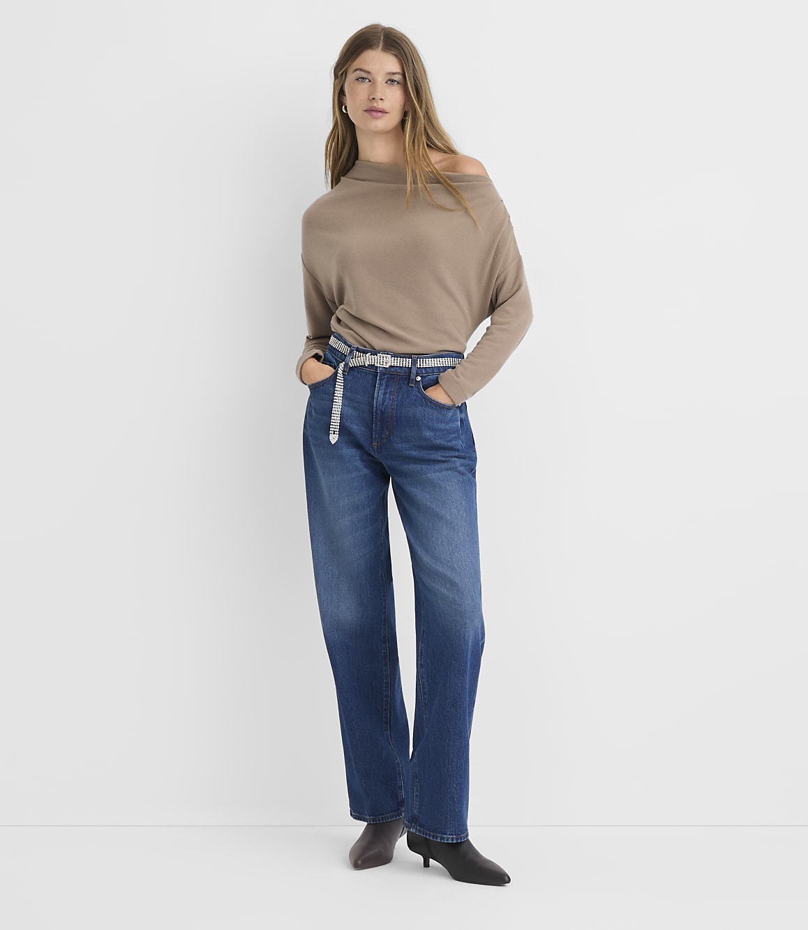 A woman in a taupe off-the-shoulder sweater and dark wash jeans stands against a plain white background.