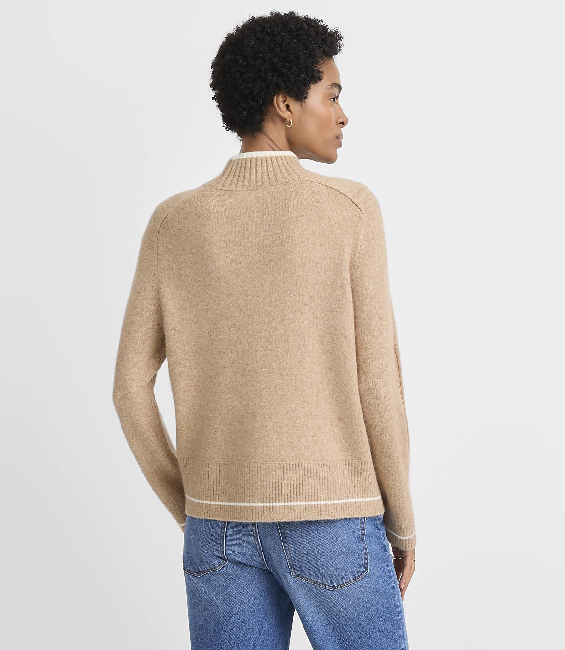 A woman in a tan mock-neck sweater and blue jeans stands against a plain, light gray background.