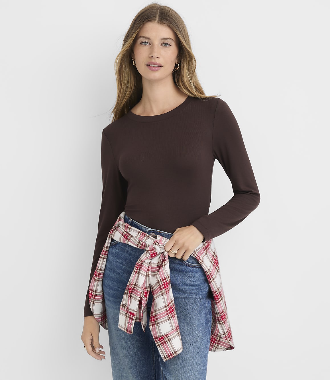 A woman in a brown long-sleeved shirt and jeans stands against a plain white background, with a red and white plaid shirt tied around her waist.