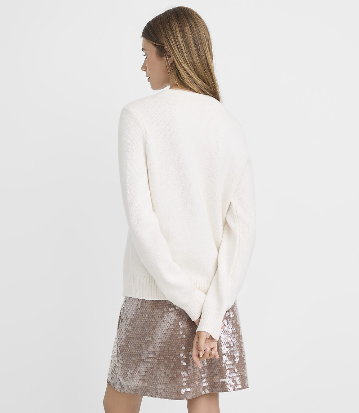 A woman in a cream sweater and sequined skirt stands against a plain white background.