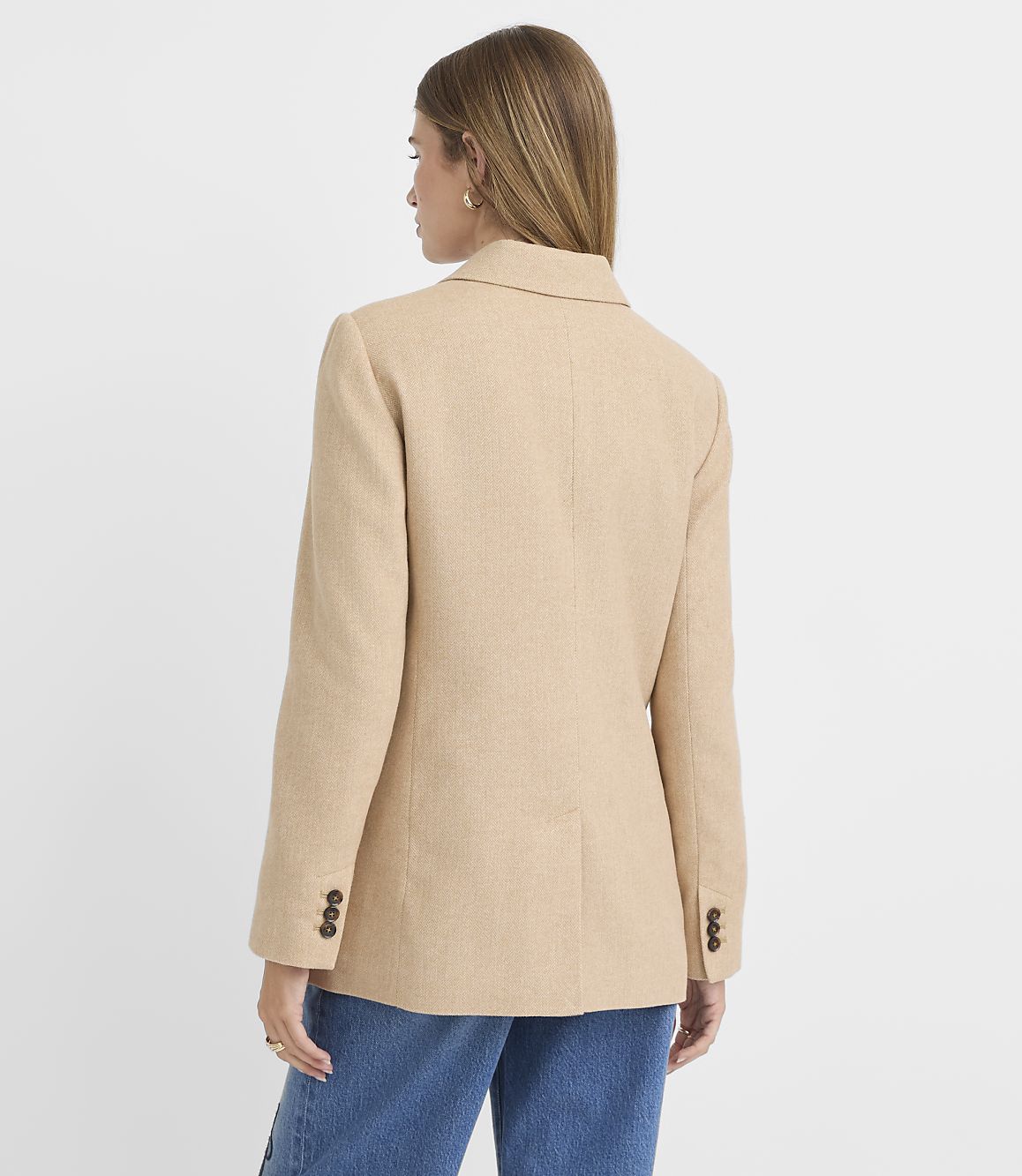 A woman in a tan blazer and blue jeans stands against a plain white background.
