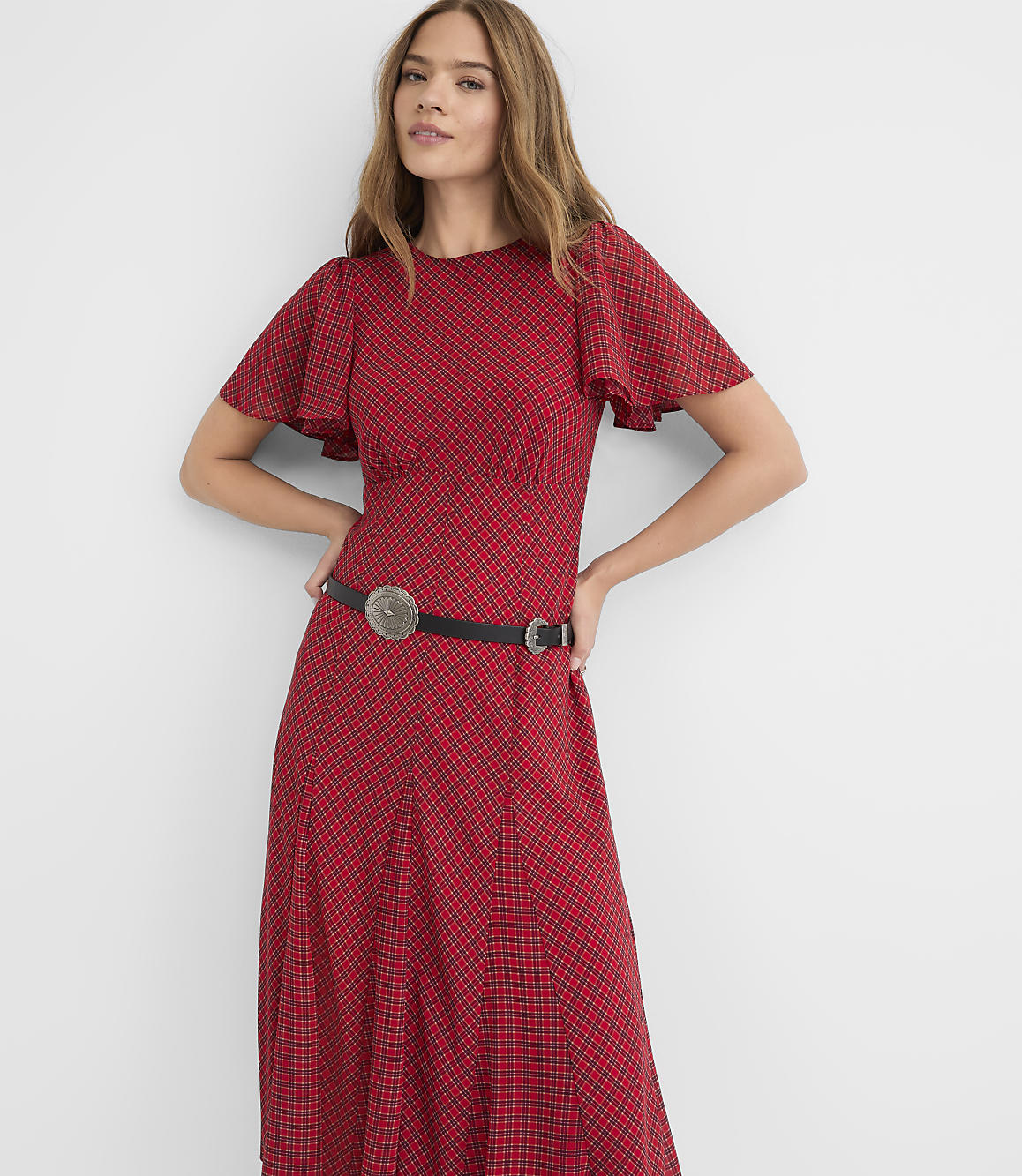 A woman in a red patterned dress with flutter sleeves and a western-style belt stands against a plain white background.