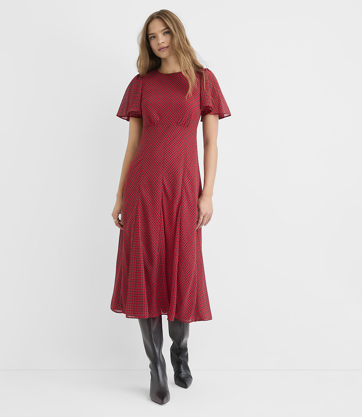 A woman in a red polka dot midi dress and black boots stands against a plain white background.