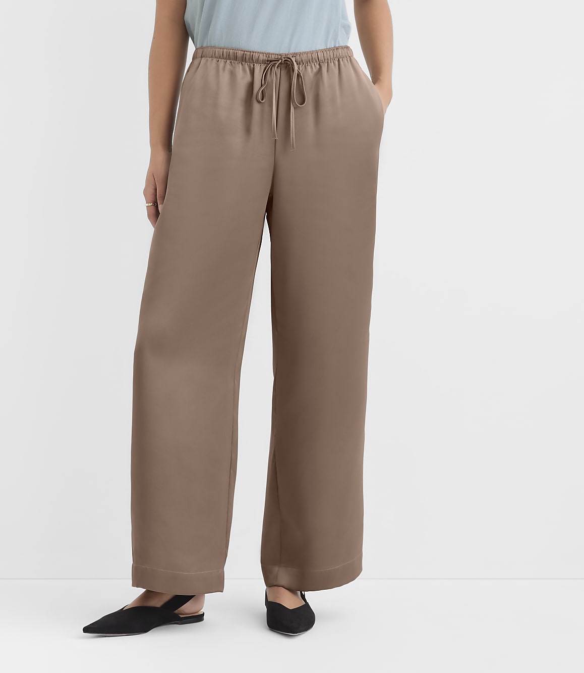 A person wearing loose, taupe, wide-leg pants with a drawstring waist and black pointed-toe flats stands against a plain white background.