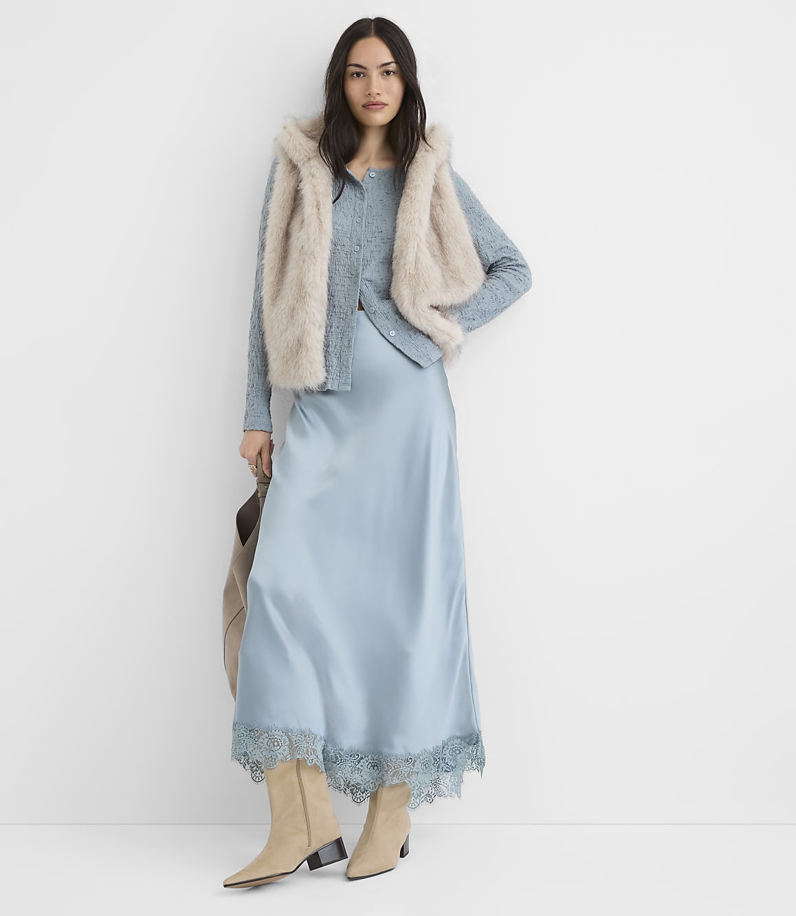 A woman in a light blue satin slip dress and a fuzzy vest stands against a plain white background.