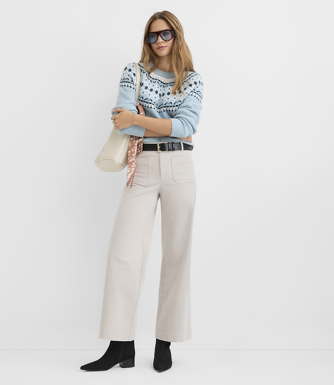 A woman in a light blue patterned sweater and cream-colored wide-leg pants stands against a plain white background.