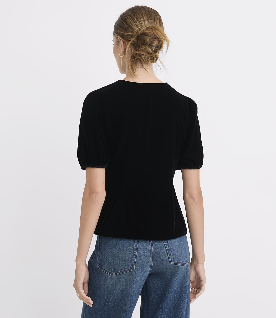 A woman in a black velvet top and blue jeans stands against a plain white background.