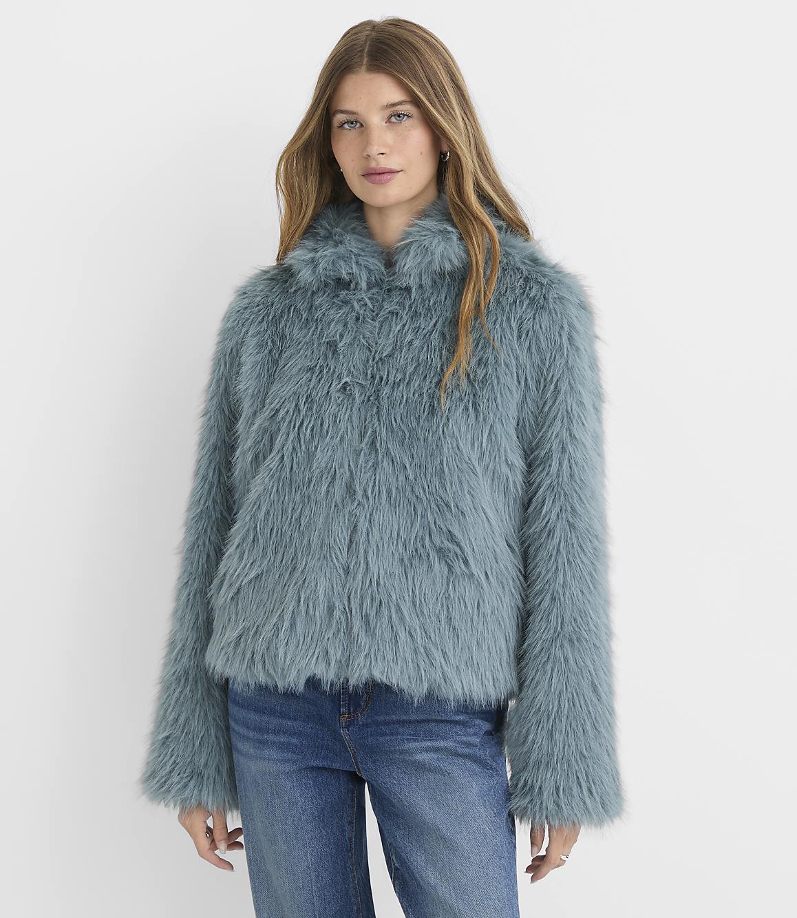 A woman in a teal faux fur jacket and blue jeans stands against a plain white background.