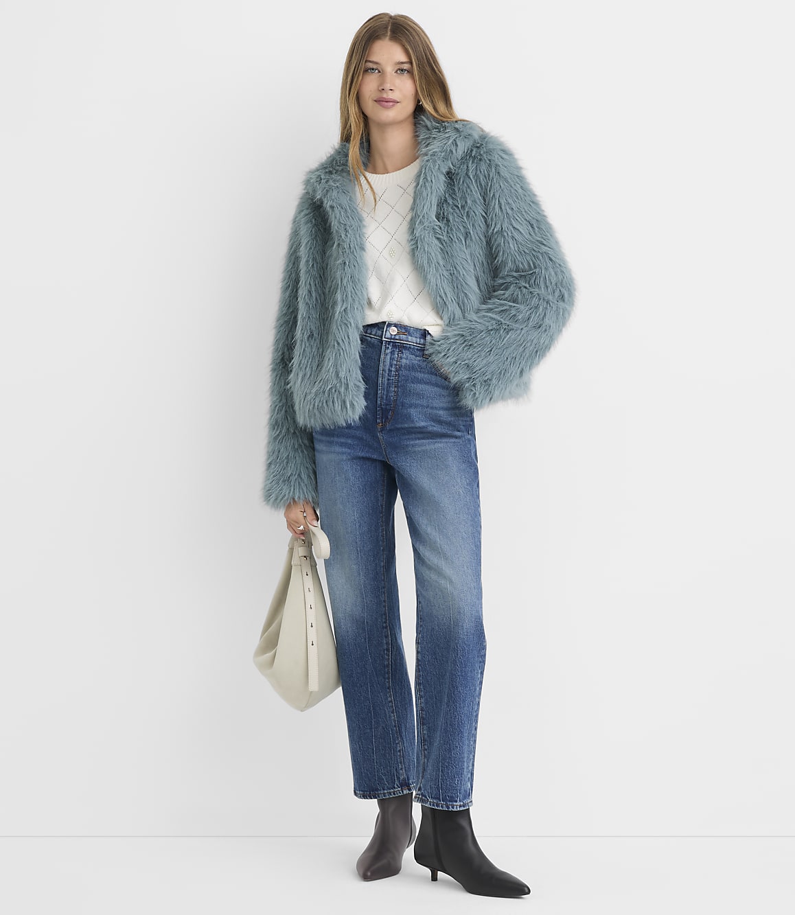 A woman in a fluffy blue jacket and jeans stands against a plain white background, holding a cream-colored bag.
