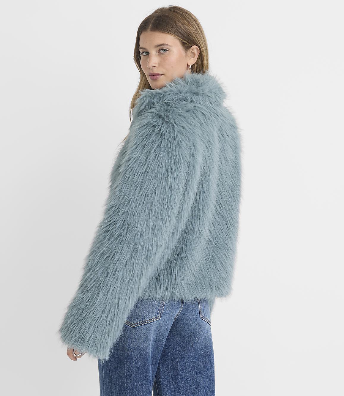 A woman in a fluffy blue jacket and jeans stands against a plain white background.