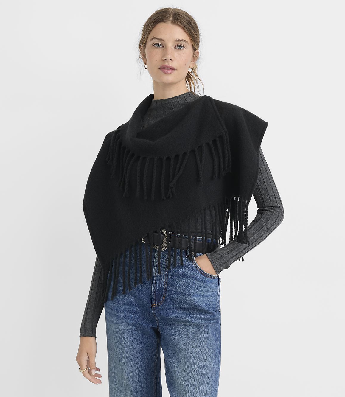 A woman in a black fringed shawl and jeans stands against a plain gray background.