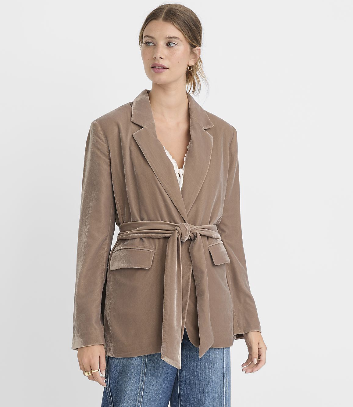A woman in a taupe velvet blazer cinched with a matching belt stands against a plain white background.