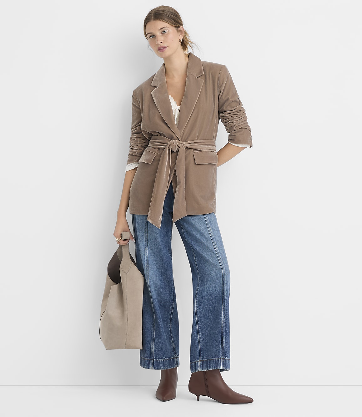 A woman in a brown belted blazer and wide-leg jeans stands against a plain white background, holding a taupe tote bag.