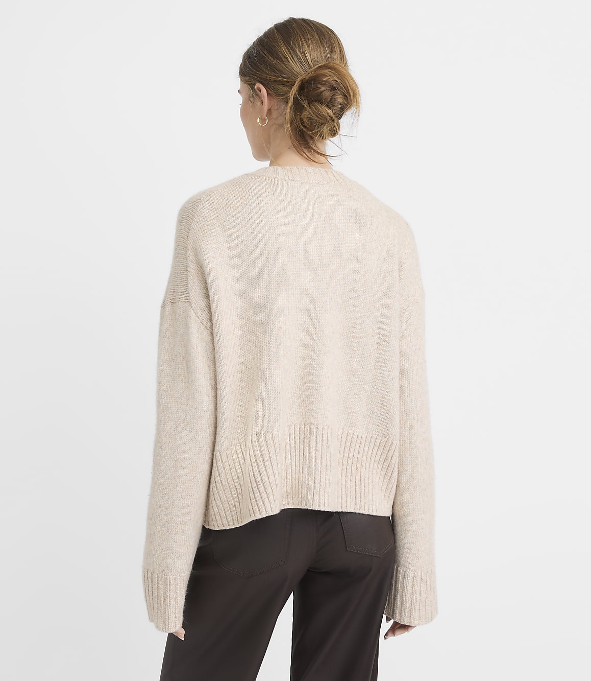 A woman in a beige sweater and dark pants stands against a plain white background.
