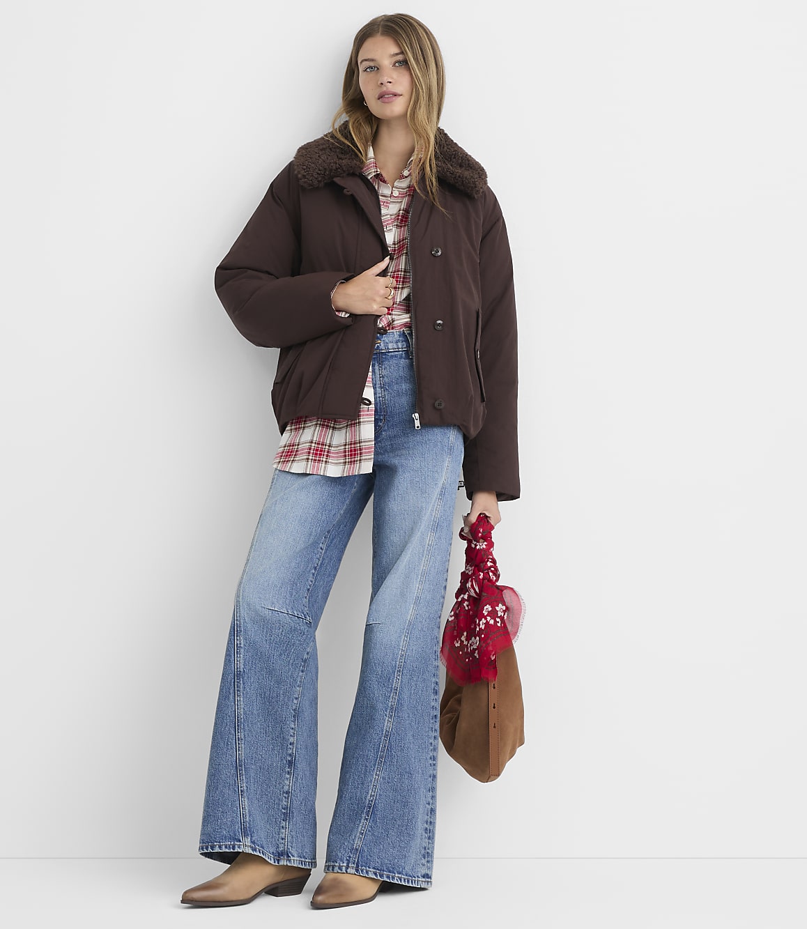 A woman in a brown puffer jacket and flared jeans stands against a plain white background, holding a brown bag with a red scarf.