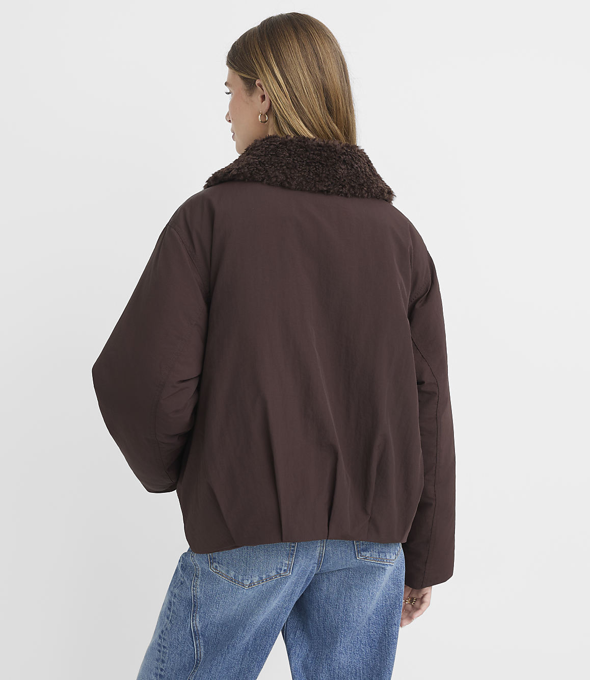 A woman in a brown puffer jacket with a fur collar and blue jeans stands against a plain white background.