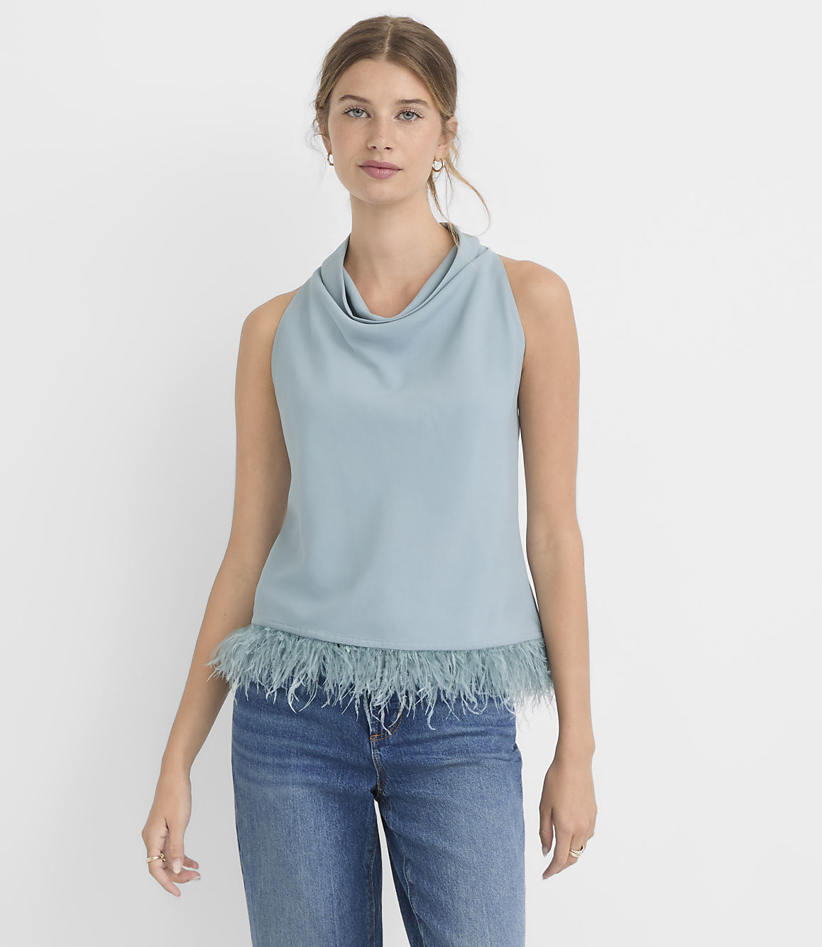 A woman in a blue cowl-neck sleeveless top with a feathered hem and blue jeans stands against a plain white background.