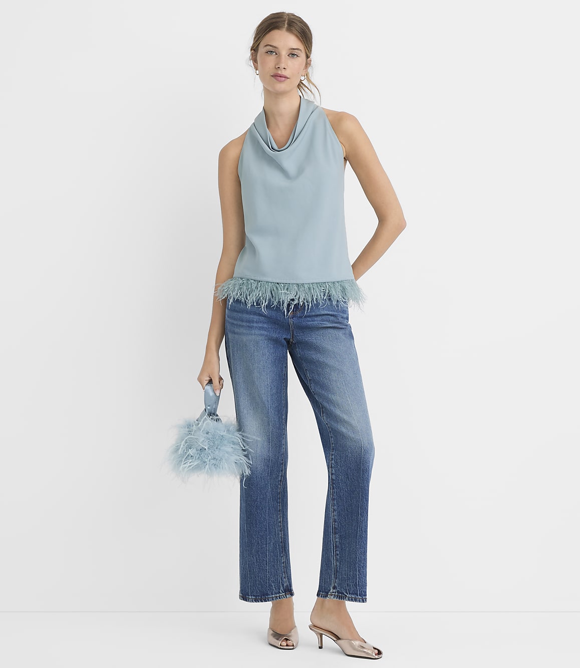 A woman in a light blue cowl-neck top with feathered trim and blue jeans stands against a plain white background, holding a matching feathered clutch.