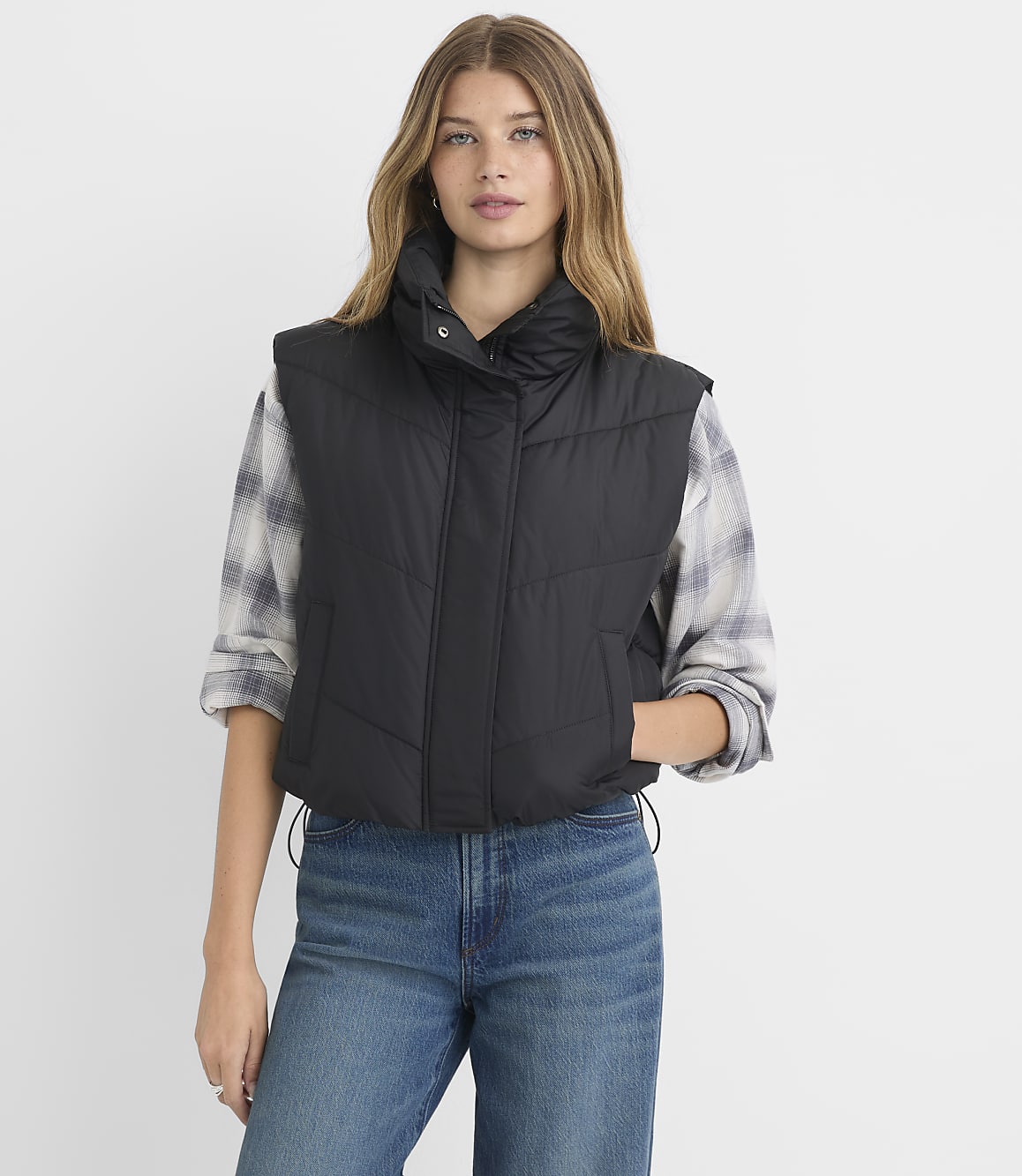 A woman in a black puffer vest and plaid shirt stands against a plain white background.