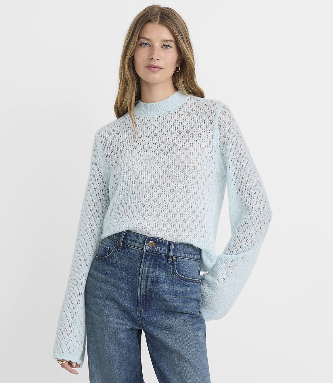 A woman in a light blue, open-knit sweater and blue jeans stands against a plain, light blue background.