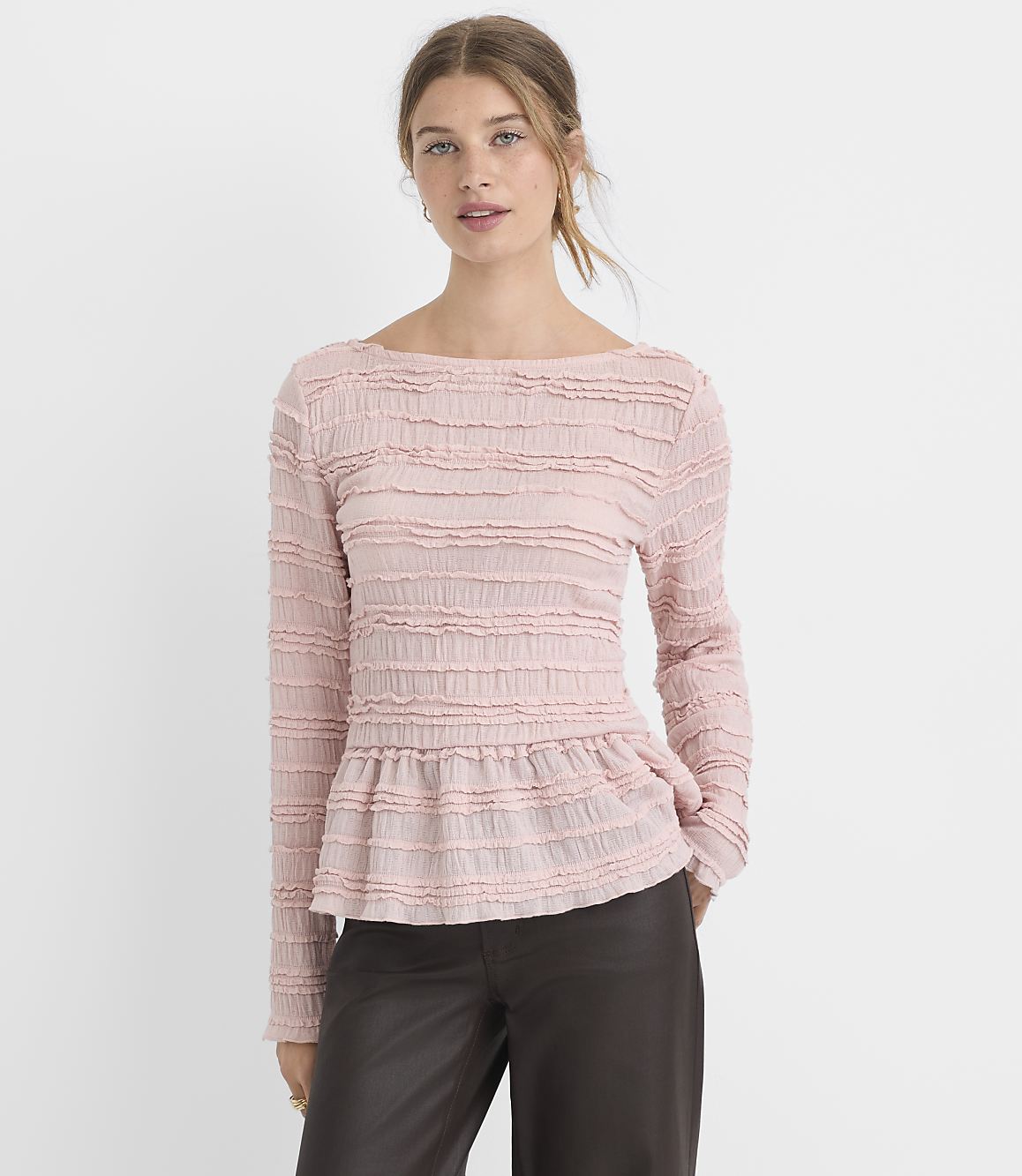 A woman in a pink textured peplum top and dark pants stands against a plain white background.