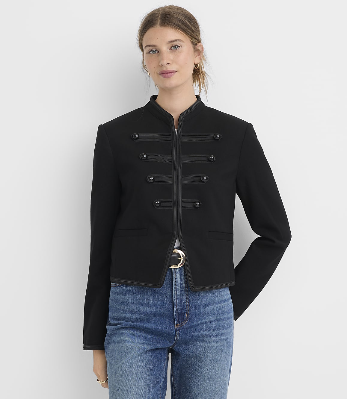 A woman in a black military-style jacket and blue jeans stands against a plain white background.