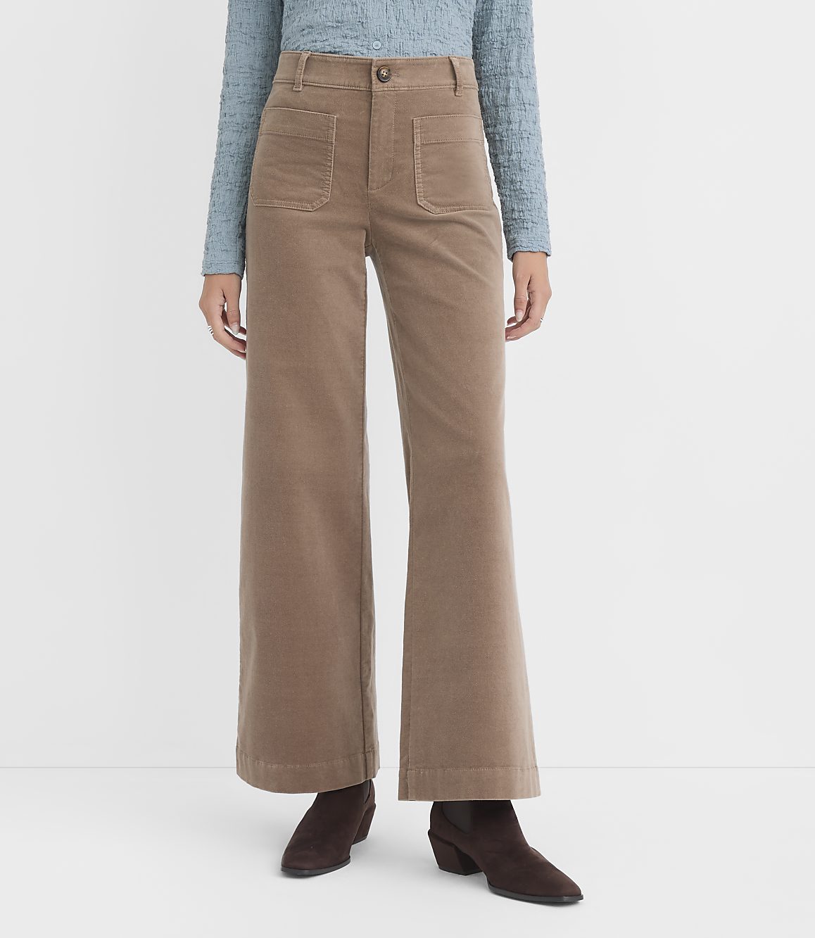 A person stands in front of a white background wearing a blue textured shirt and tan corduroy wide-leg pants with brown boots.
