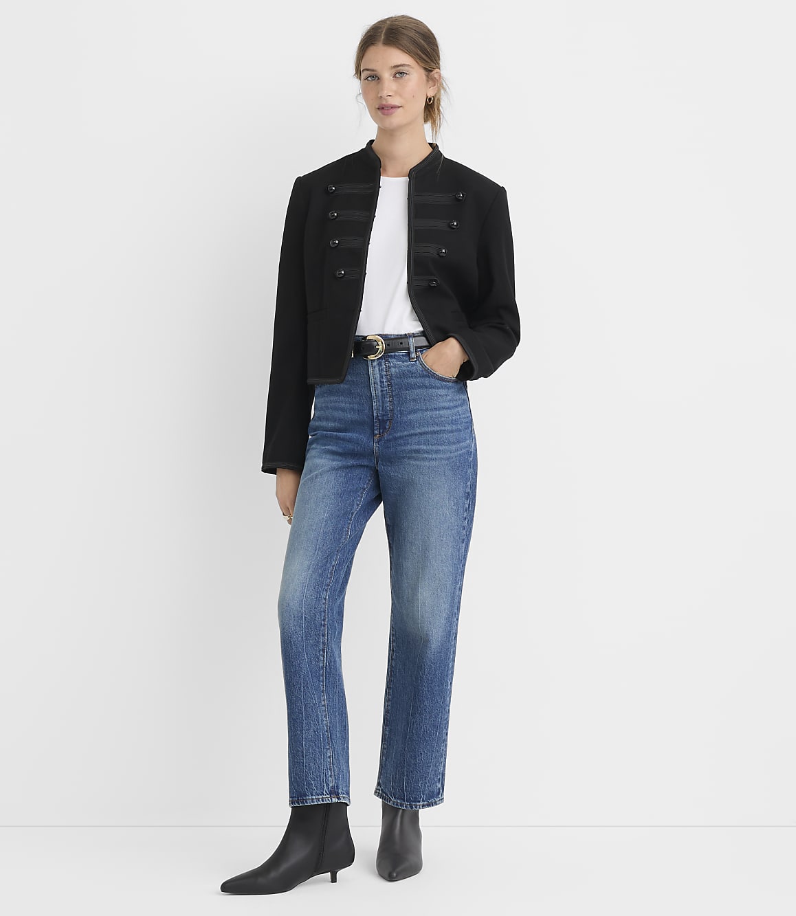 A woman in a black military-style jacket and blue jeans stands against a plain white background.