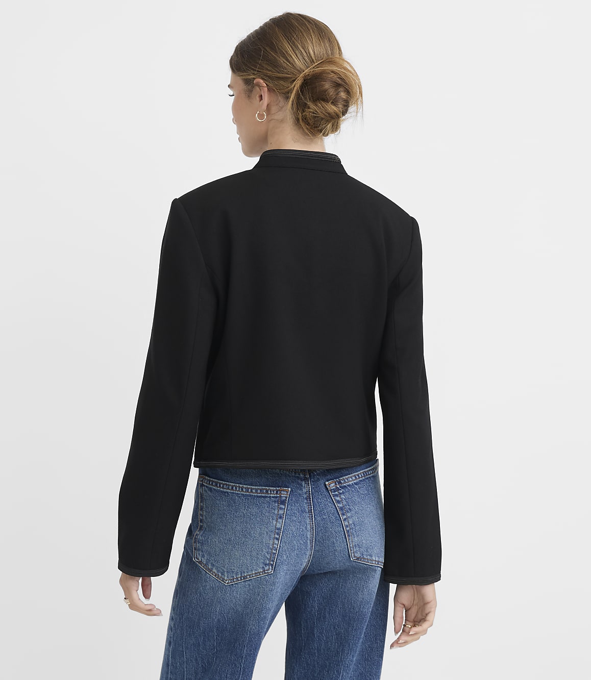 A woman in a black jacket and blue jeans stands against a plain white background.