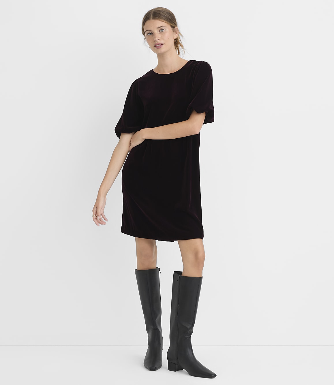 A woman in a dark velvet dress and black boots stands against a plain white background.