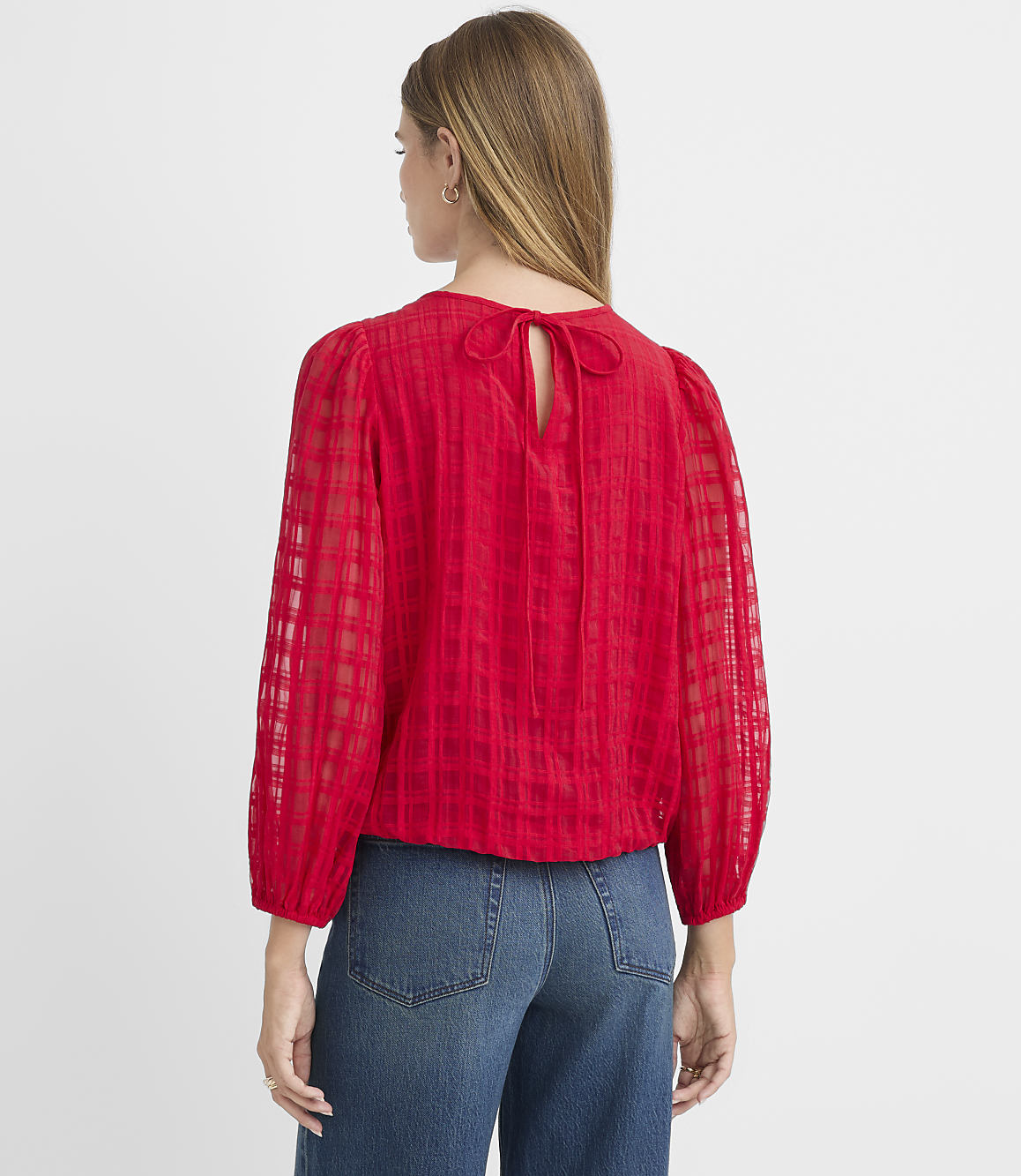 A woman in a red checkered blouse with a bow at the back stands against a plain white background.