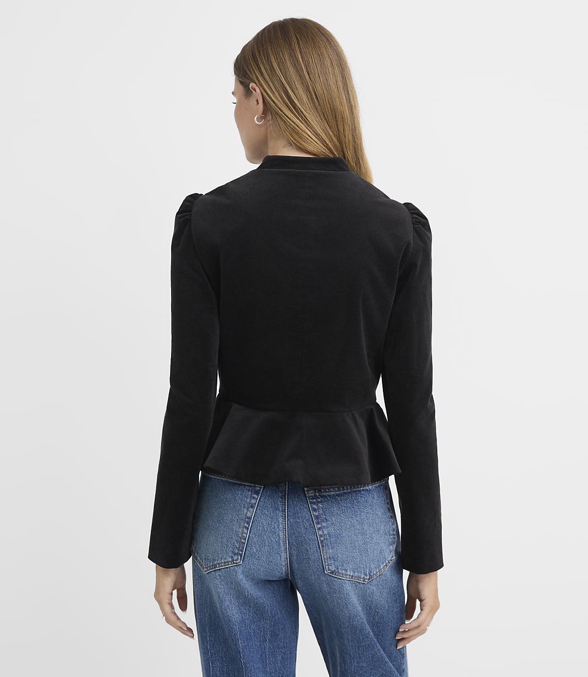 A woman in a black peplum jacket and blue jeans stands against a plain gray background.