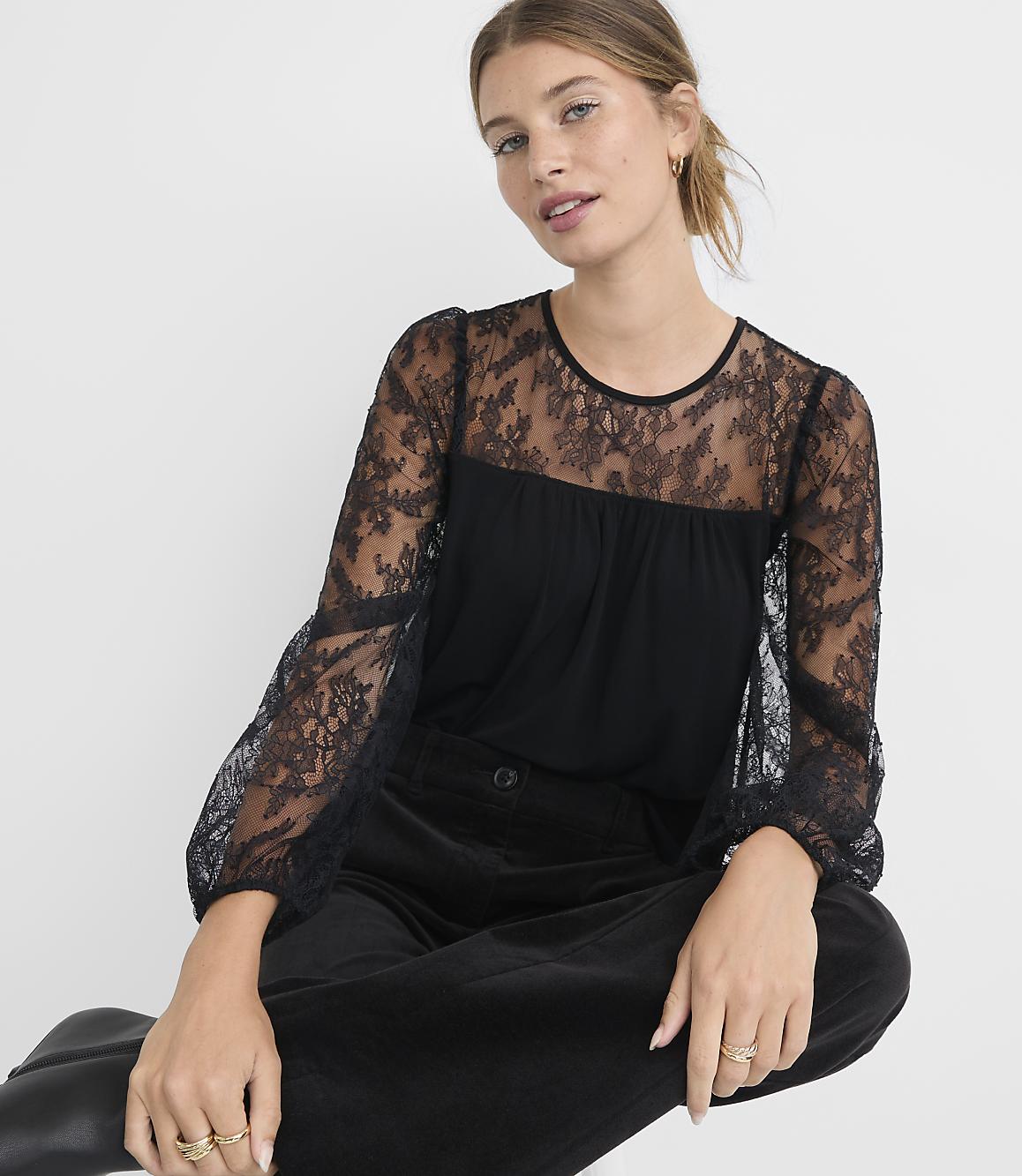 A woman in a black lace-sleeved top and dark pants sits against a plain white background.