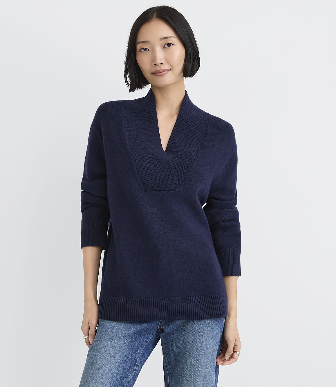 A woman wearing a navy blue V-neck sweater and blue jeans stands against a plain white background.