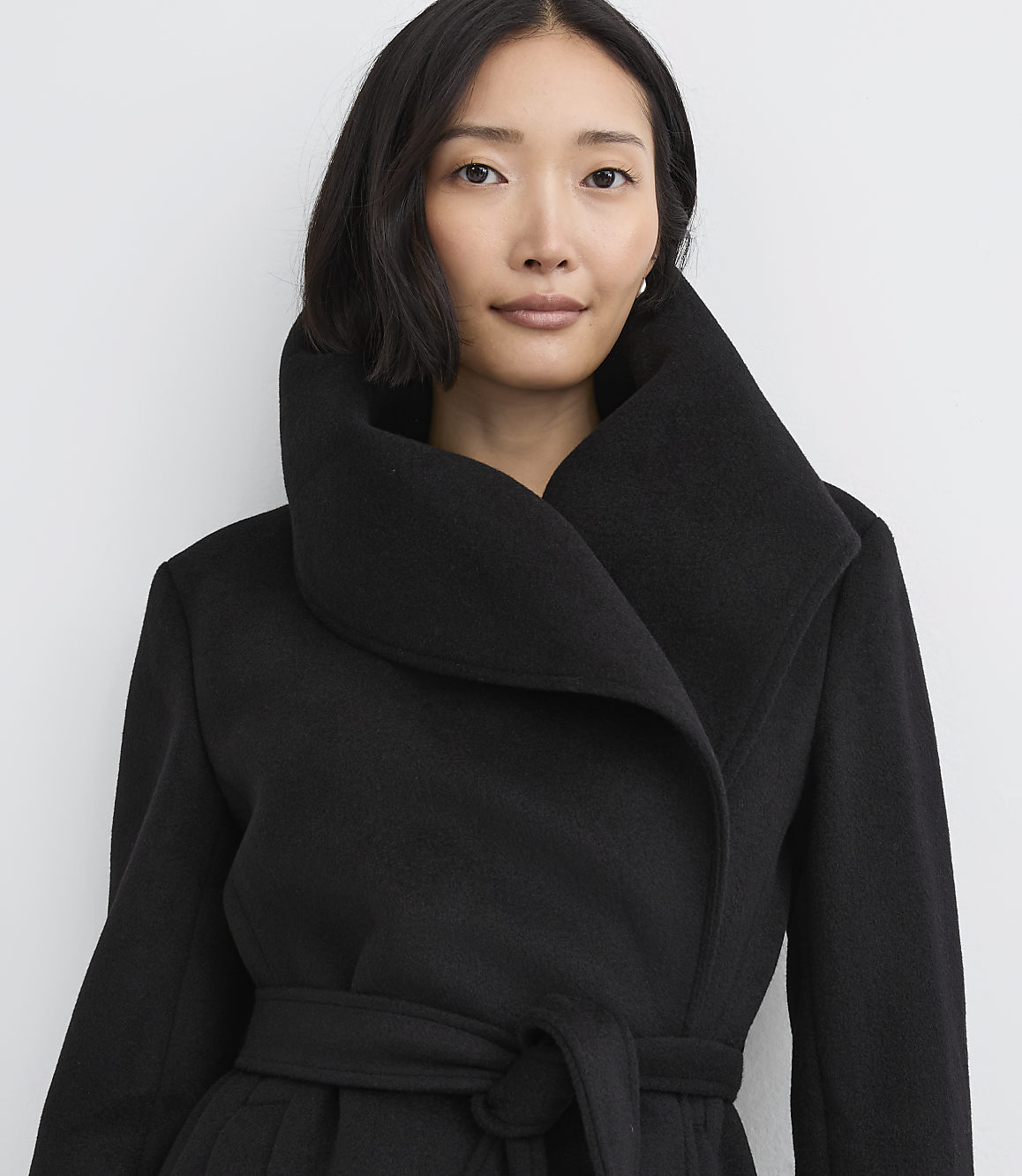 A woman with dark hair wearing a black belted coat stands against a plain white background.