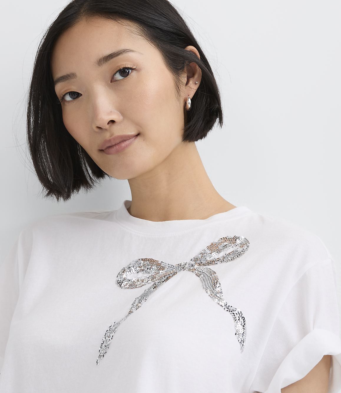 A woman wearing a white t-shirt with a silver sequined bow stands against a plain white background.