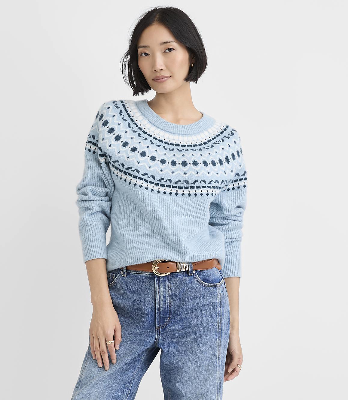A woman wearing a light blue patterned sweater and blue jeans stands against a plain white background.