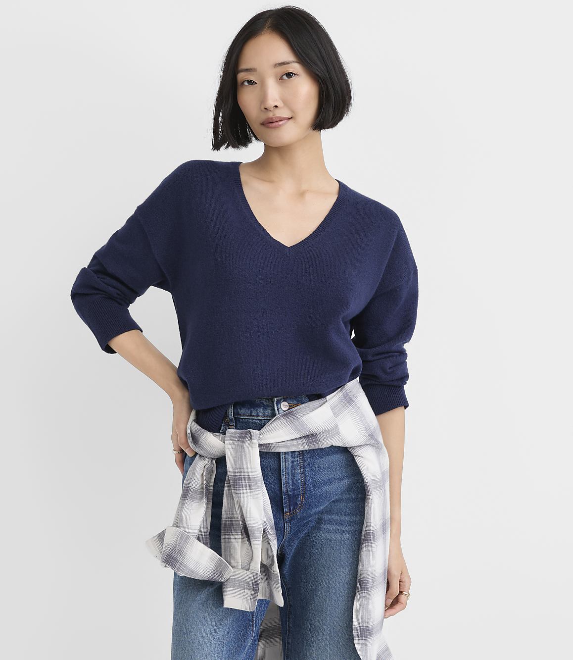 A woman wearing a navy blue V-neck sweater and blue jeans with a plaid shirt tied around her waist stands against a plain white background.