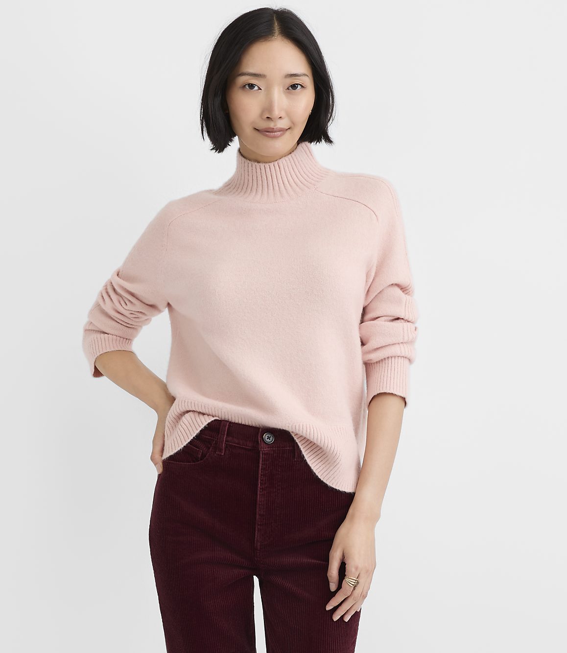 A woman wearing a light pink turtleneck sweater and burgundy corduroy pants stands against a plain white background.